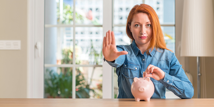 Redhead woman saves money in piggy bank at home with open hand doing stop sign